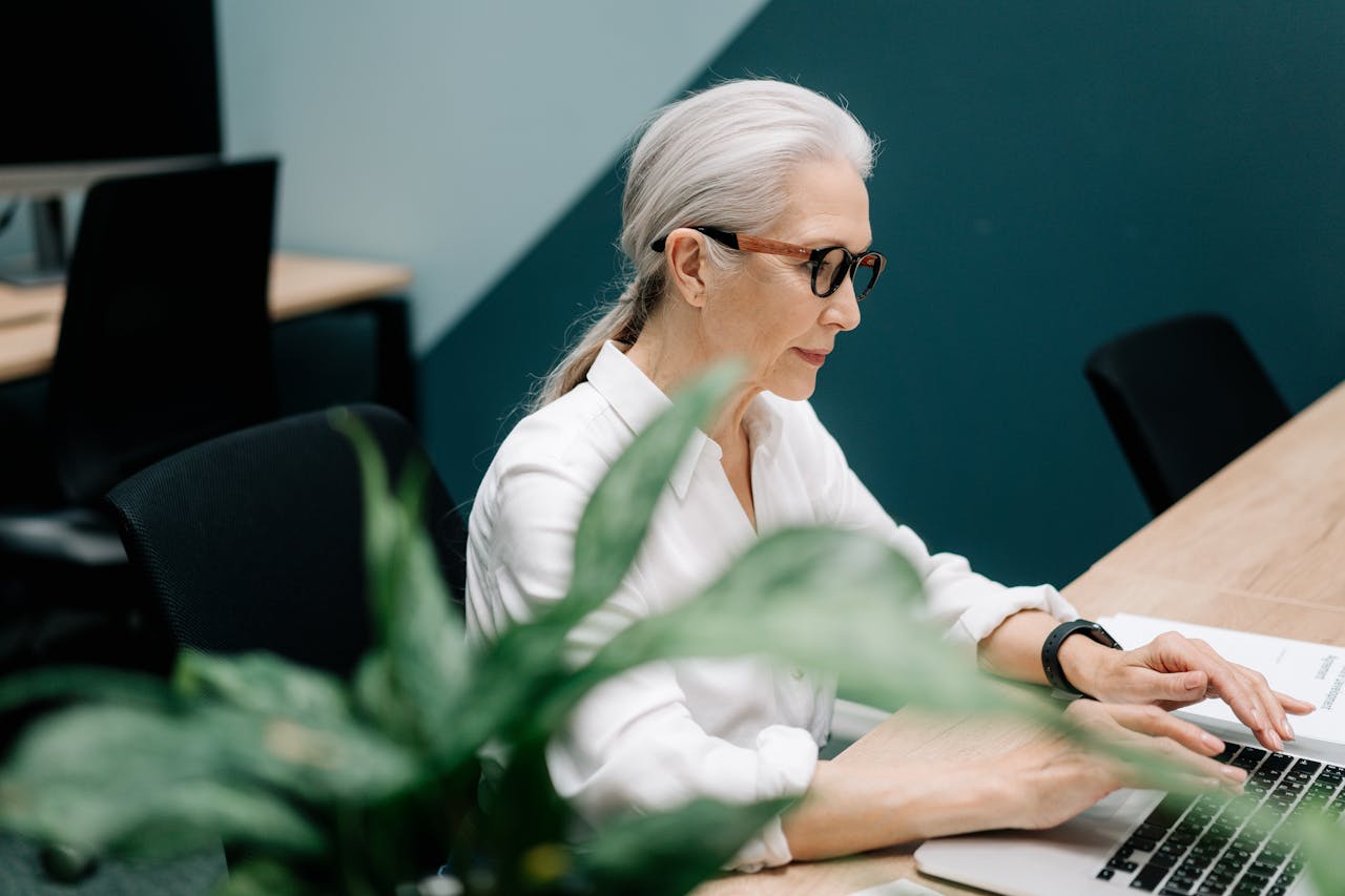 Confident senior woman with glasses working on a laptop in a modern office setting.